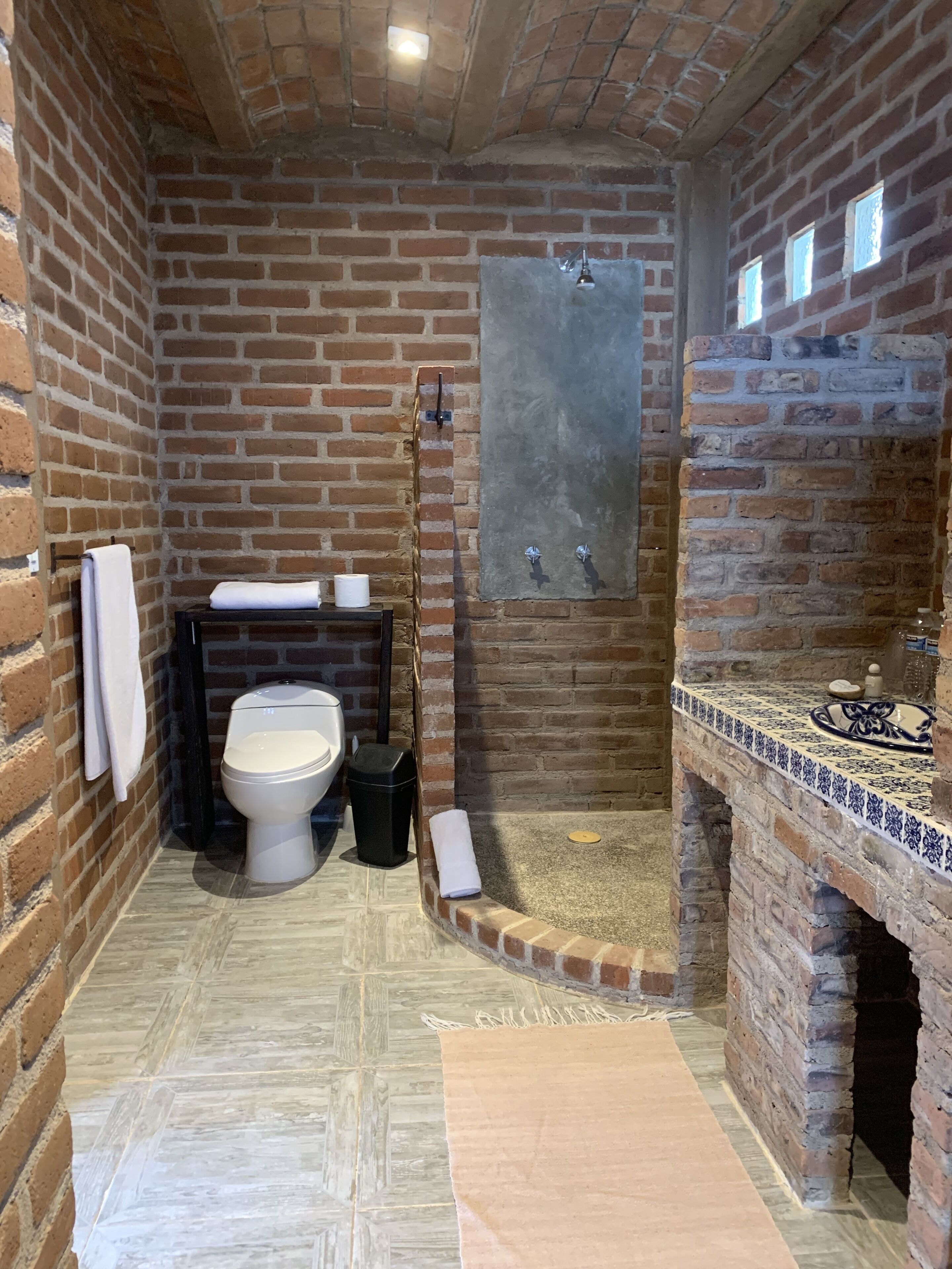 A bathroom with a brick wall, a toilet, a sink with a tiled countertop, and a shower area.