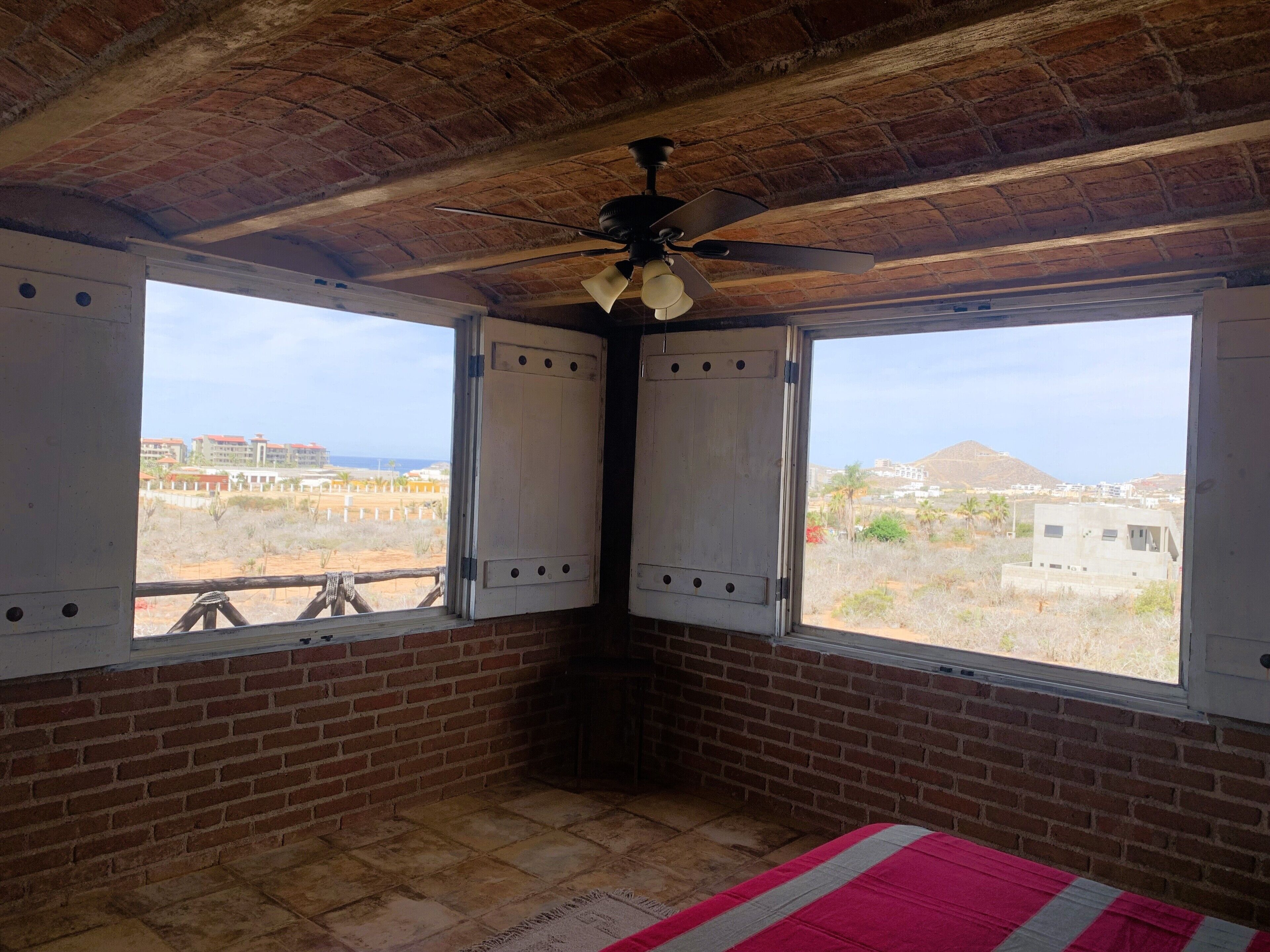A room with a brick floor, two windows with white shutters, a ceiling fan, and a view of buildings and a hill.