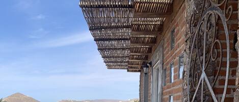 Una terraza de madera con barandilla de metal, un muro de ladrillo con detalles metálicos decorativos y vistas a edificios y colinas distantes.