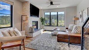 A modern living room with a stone fireplace, large windows, a ceiling fan, and a view of a forest. - Downtown WP Loft 49 I Rooftop Hot Tub & Lounge I Great Views I Bunk Room I Discounted Attractions (Winter Park)