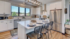 A modern kitchen with white cabinets, a large island with bar stools, stainless steel appliances, and a view of the outdoors. - Downtown WP Loft 49 I Rooftop Hot Tub & Lounge I Great Views I Bunk Room I Discounted Attractions (Winter Park)
