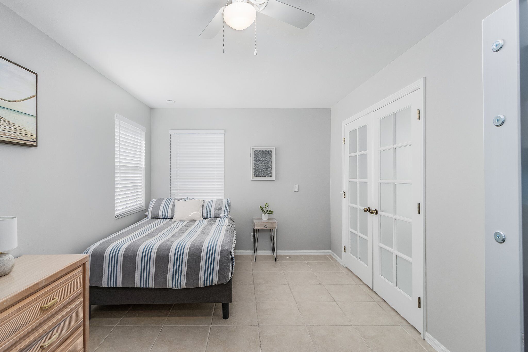 A bedroom with a bed, bedside table, lamp, window blinds, a ceiling fan, a door, and a small table with a plant.