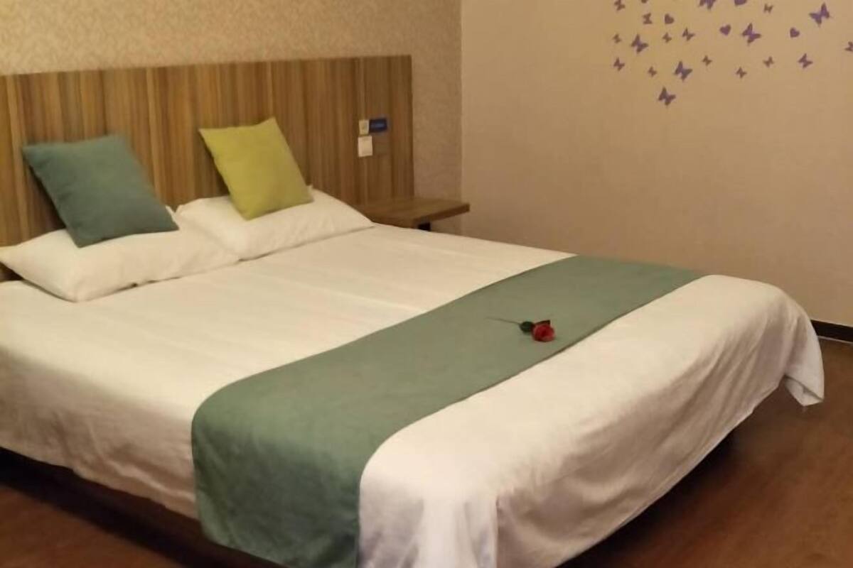 Un lit bien fait, recouvert d’une couvre-lit verte et blanche, avec deux oreillers et une seule rose posée sur le lit.