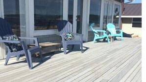 Um deck de madeira com duas cadeiras Adirondack e uma mesa, porta de entrada de vidro e vista para o oceano.