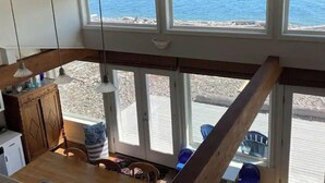 Um quarto com pé direito alto, grandes janelas com vista para a praia, vigas de madeira e um espaço de jantar com cadeiras e mesa.
