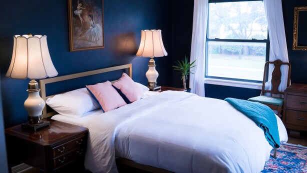 A neatly made bed with white linens and a blue throw blanket, flanked by two bedside lamps. A framed painting hangs above the bed, and a window with white curtains allows natural light into the room. - A Tasteful Historic 4 Bedroom Home (Oklahoma City)