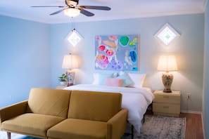 Une chambre à coucher avec un lit, deux lampes de chevet, un ventilateur de plafond, un tableau abstrait aux couleurs vives et un canapé de couleur moutarde.