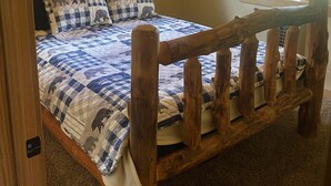 Een houten bed met een geruit sprei, een hoofdbord van boomstammen en een raam met uitzicht op bomen.