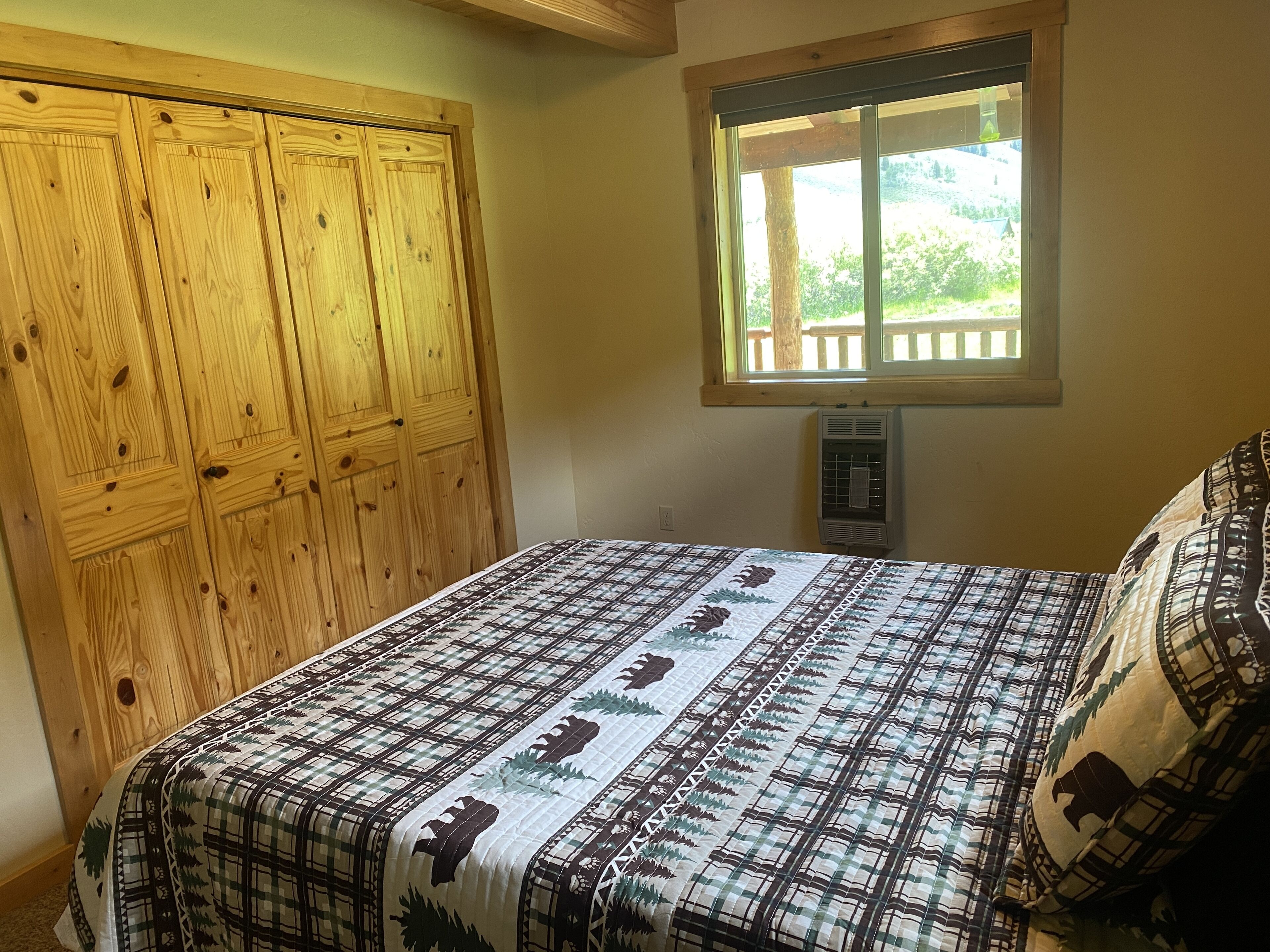 Une chambre avec une armoire en bois, un lit recouvert d’une couvre-lit à motifs, une fenêtre donnant sur la verdure et un radiateur fixé au mur.