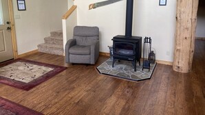 Een kamer met een grijze fauteuil, een houtkachel, een trap en een welkomstbord.