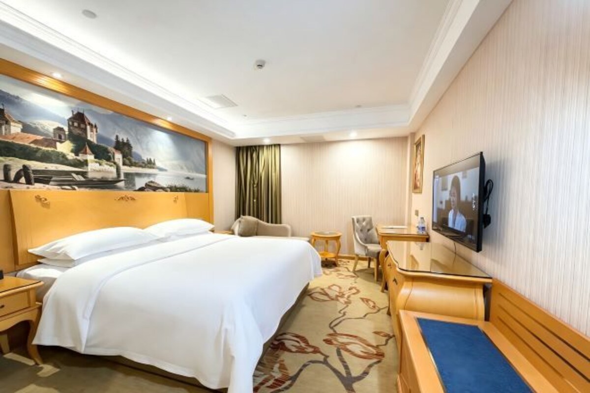 A hotel room with a bed, a desk, a chair, a television, and a painting on the wall.