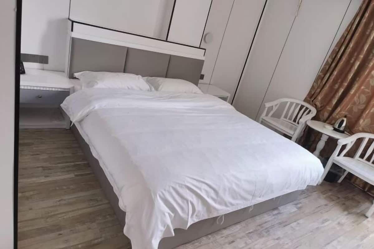 A single bed with a headboard, a small table, and two white chairs in a room with wooden flooring and white walls.