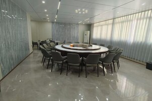 Un salón de conferencias con una mesa redonda rodeada de sillas, una pequeña mesa con algunos objetos y una ventana con persianas.