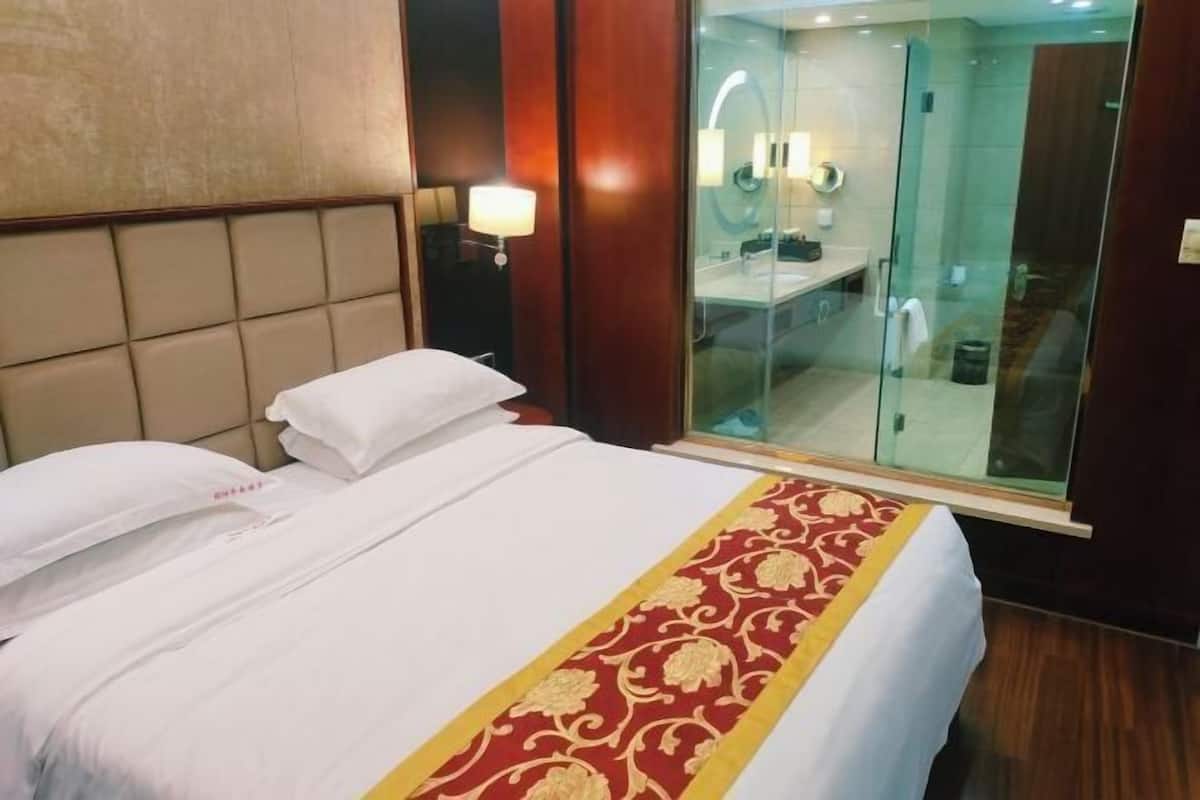 A hotel room with a bed, a nightstand, a lamp, and a glass-enclosed bathroom.