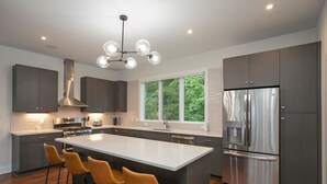 Une cuisine moderne dotée d’un grand îlot central, de meubles de rangement aux couleurs sombres, d’appareils électroménagers en acier inoxydable et offrant une vue sur la verdure environnante.