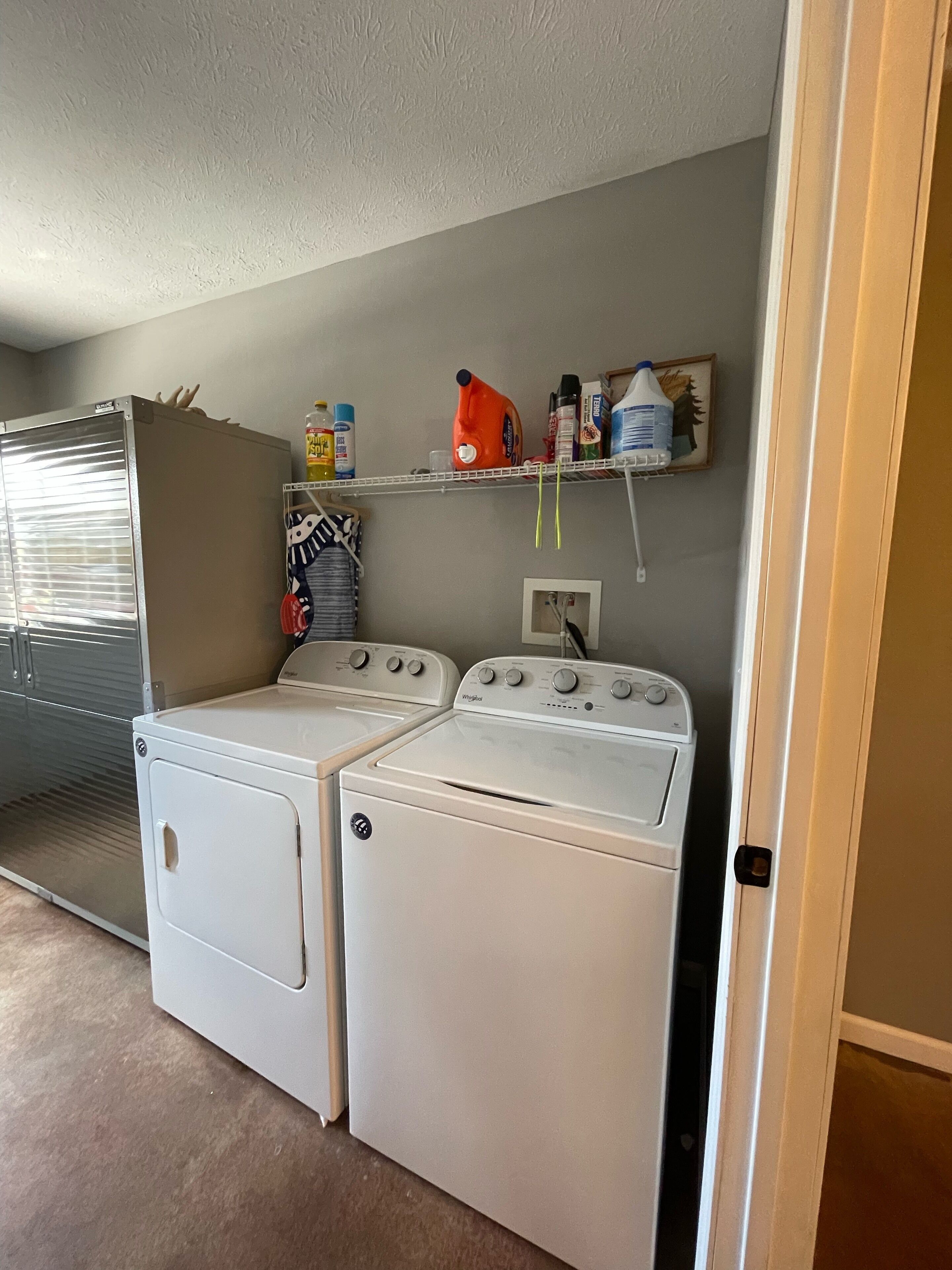 A laundry room with two white front-loading washers and dryers, a shelf with cleaning supplies, and a window with blinds.