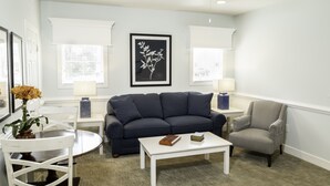 Ein Wohnzimmer mit einem dunkelblauen Sofa, einem weißen Couchtisch, einem grauen Sessel, einem kleinen runden Tisch mit Blumenvase und zwei Beistelltischen mit Lampen.