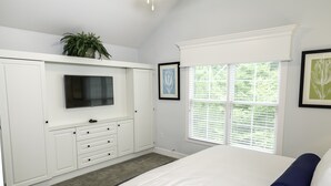 Ein Schlafzimmer mit einem Bett, einem an einer weißen Kommode befestigten Fernseher, einem Fenster mit Jalousien und gerahmten Bildern an der Wand.