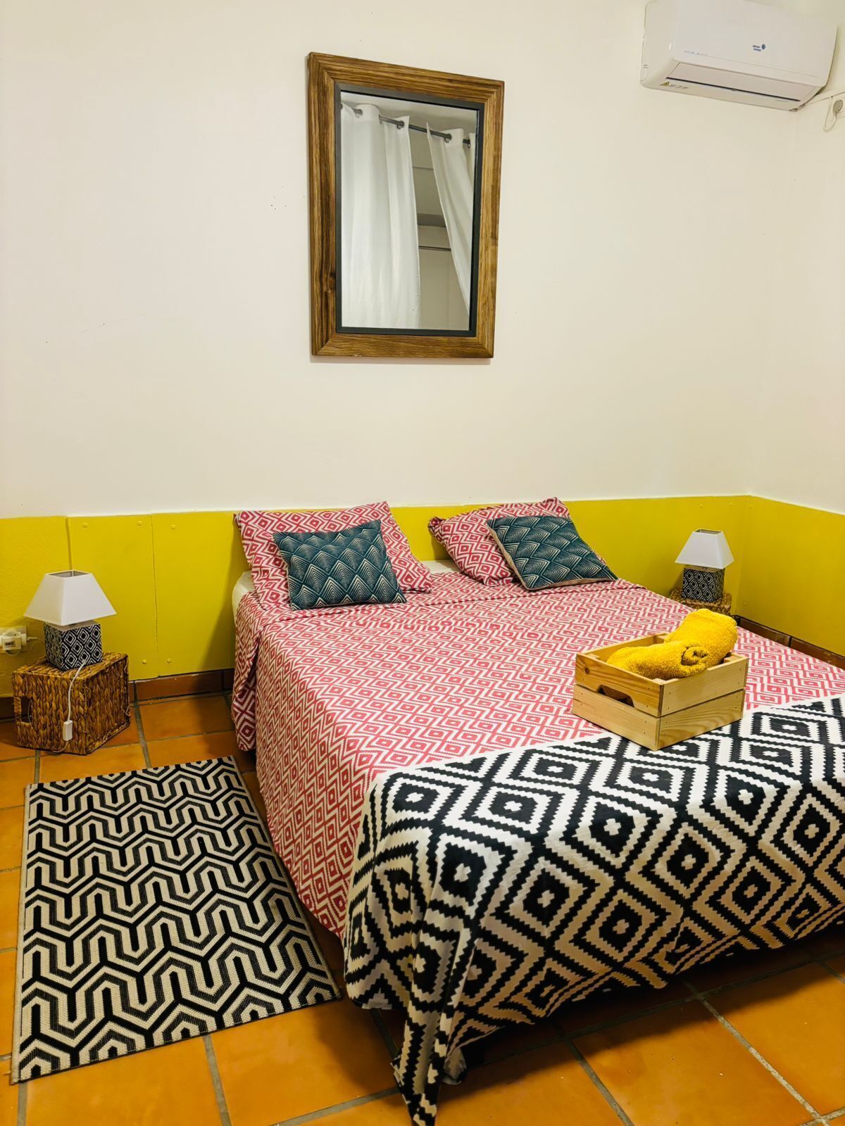 Une petite pièce aux couleurs vives, avec un lit, deux tables de chevet, un miroir et une petite fenêtre.