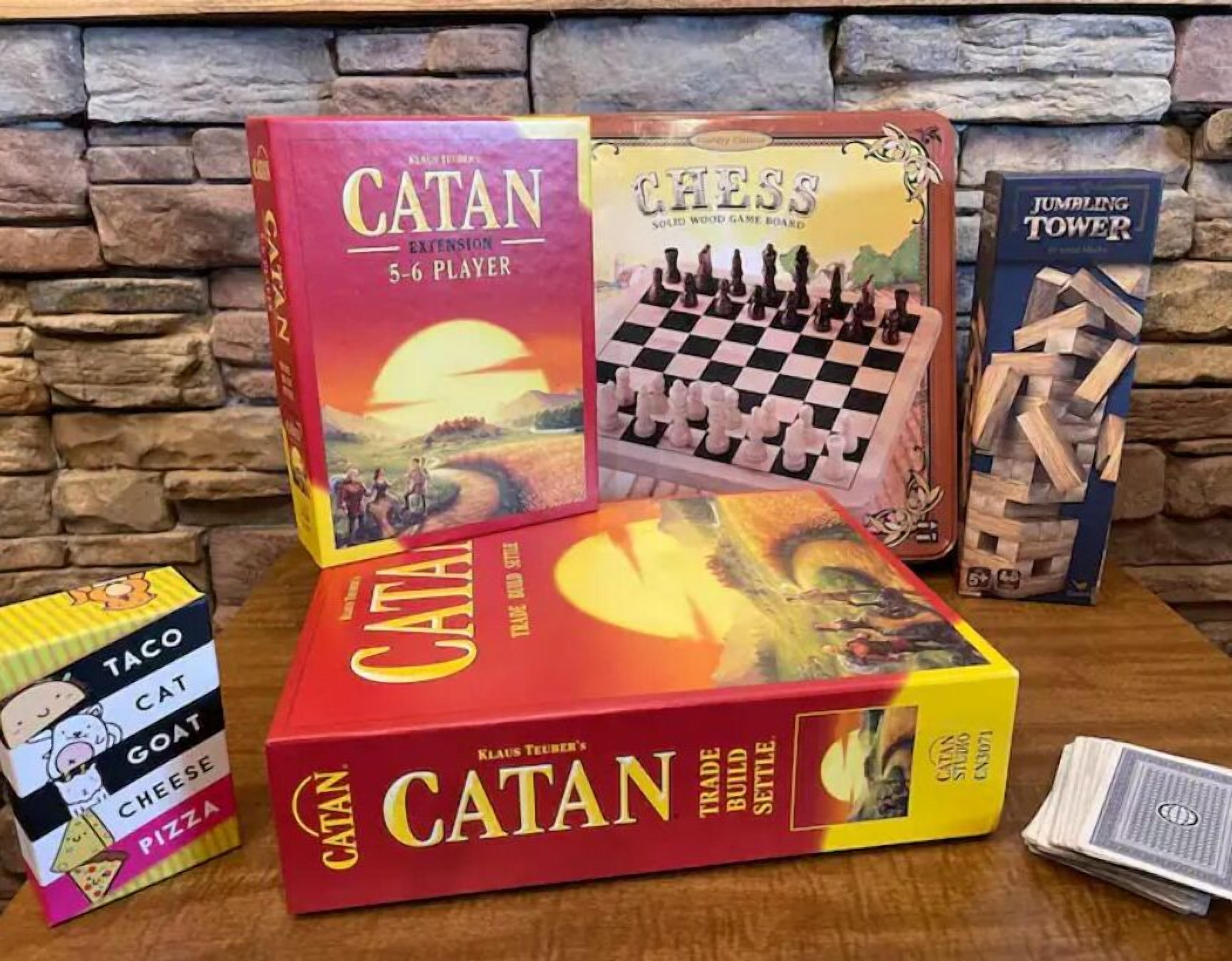 Een verzameling gezelschapsspellen, waaronder Catan, Schaken en JUMPING TOWER, op een houten tafel met een achtergrond van een stenen muur.