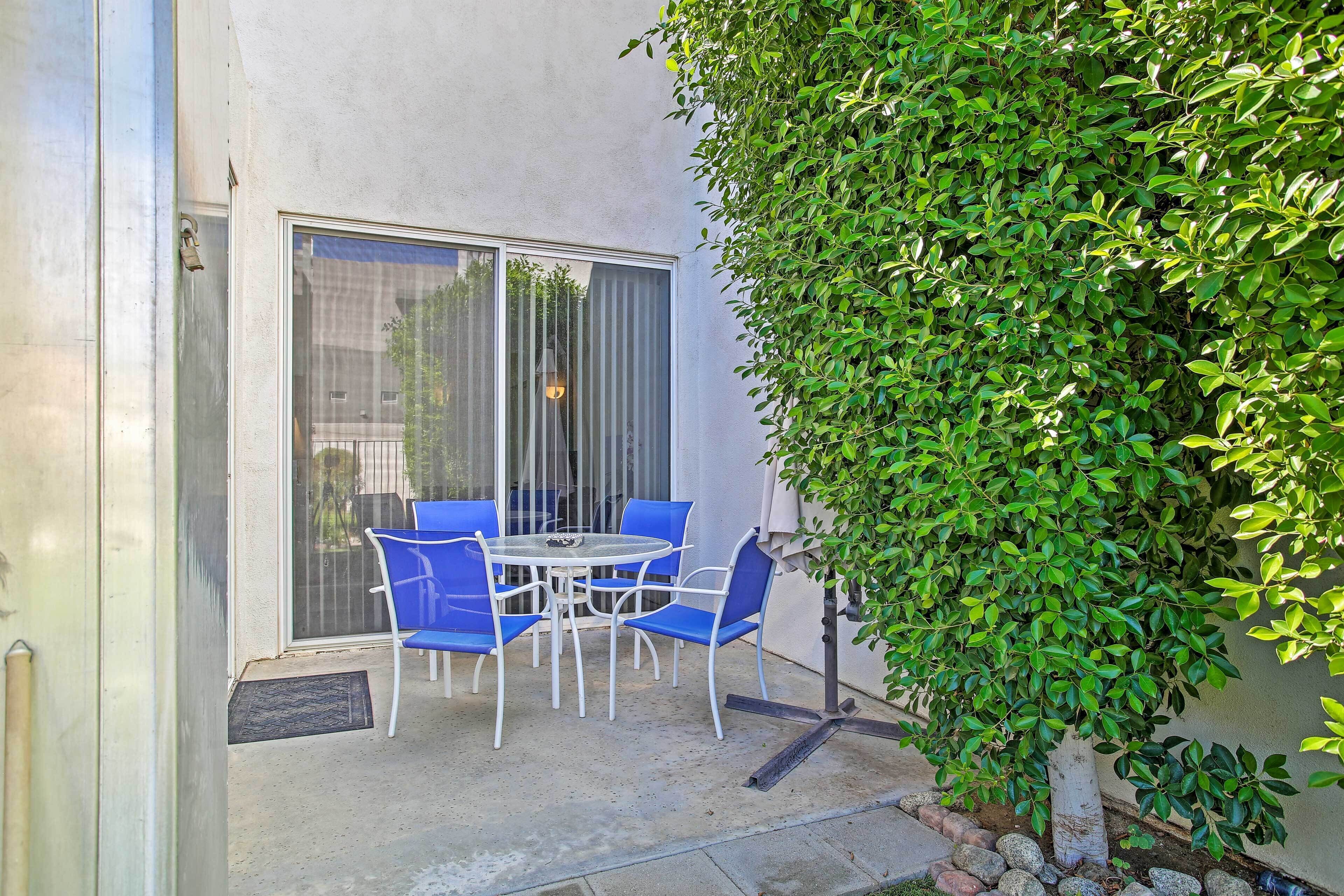 A patio with a table and blue chairs, a sliding glass door, and a green hedge.