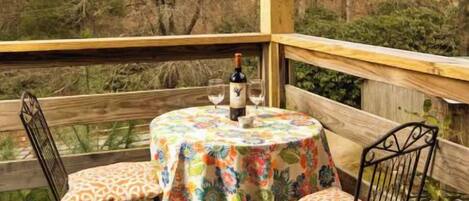 Een houten terras met een ronde tafel en twee stoelen, een fles en twee glazen op tafel.
