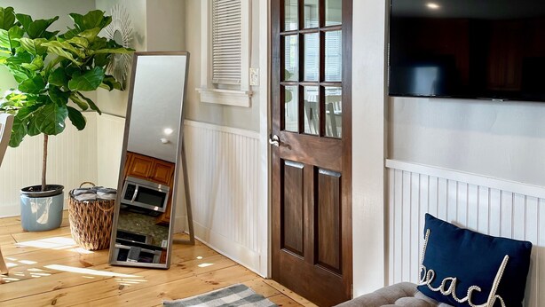 Un couloir avec un plancher en bois, une grande plante en pot, un miroir, une enseigne indiquant « MAISON DE PLAGE » et un canapé avec un coussin.
