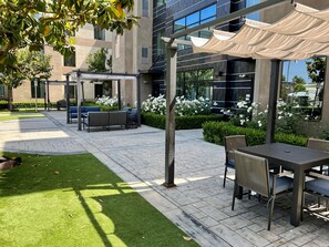 Un área de estar al aire libre con mesas y sillas, una pérgola con toldo y un edificio con grandes ventanales al fondo.