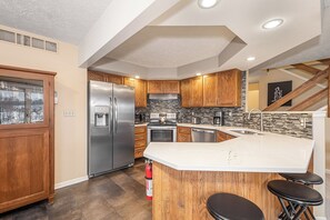 A modern kitchen with a stainless steel refrigerator, wooden cabinets, a built-in oven, and a large island with bar stools. - 180 Degrees by Taylor Made Deep Creek (Oakland)
