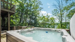 A hot tub with a stone exterior, surrounded by a wooden deck and trees, overlooking a body of water. - Sunset Pointe by Taylor Made Deep Creek (Oakland)