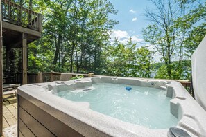 A hot tub with a stone exterior, surrounded by a wooden deck and trees, overlooking a body of water. - Sunset Pointe by Taylor Made Deep Creek (Oakland)
