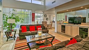 A spacious living room with a large window, a sofa with red cushions, a coffee table with a glass top, and a kitchen area with a TV.