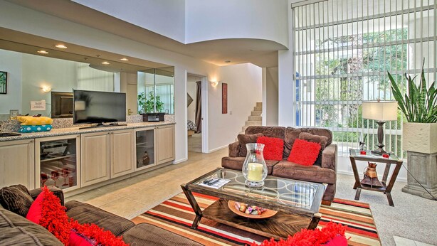 A modern living room with a glass coffee table, a brown sofa, and a kitchen area with a TV and cabinets.