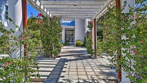 A covered walkway with white columns and a tiled floor, surrounded by flowering plants.