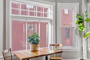 A dining area with a wooden table, chairs, a potted plant, and large windows with pink siding.