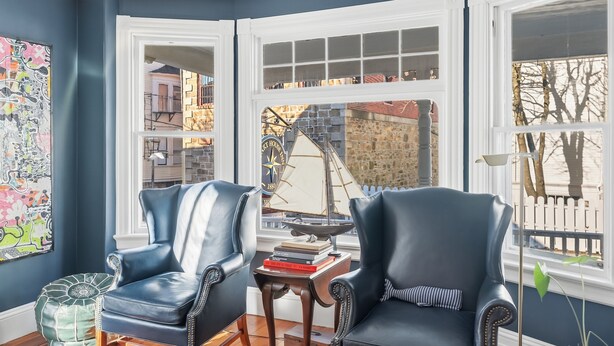 A room with two leather chairs, a small table with books, a colorful abstract painting, and a view of a brick building through a window. - Chart House Inn (Newport)