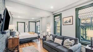 A modern bedroom with a bed, a gray sofa, a wooden chair, a desk, and a TV. - Chart House Inn (Newport)