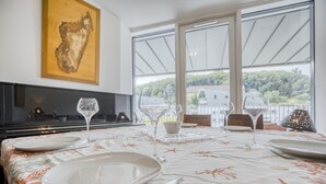Una mesa de comedor dispuesta con platos, cubiertos y copas de vino, con vistas a zonas verdes y edificios a través de amplios ventanales.