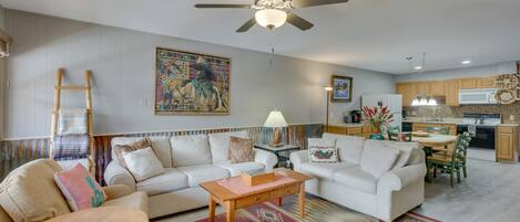 A living room with a sofa, coffee table, and a dining area with chairs. There is a ceiling fan, a lamp, and a kitchen in the background.