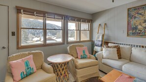 Un salon chaleureux avec un canapé beige, deux fauteuils, une petite table en bois et une grande fenêtre équipée de stores.
