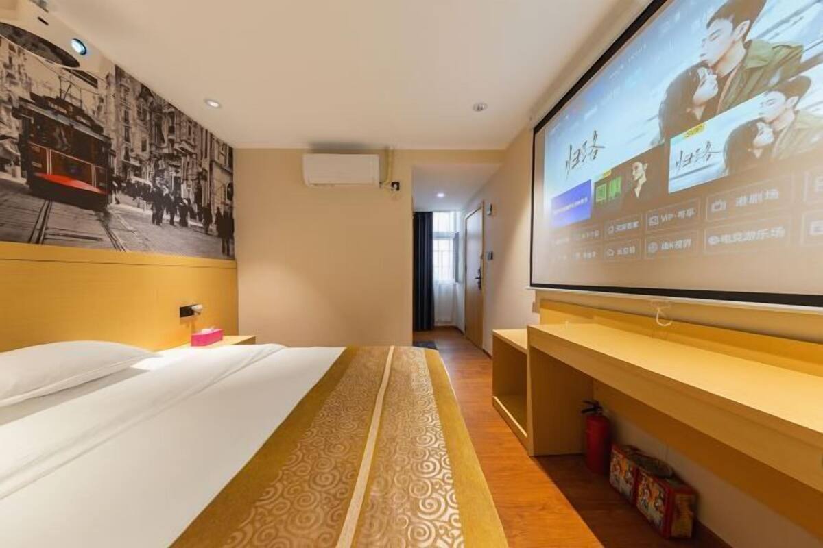 A hotel room with a bed, a TV screen displaying a movie, a wall-mounted air conditioner, and a hallway leading to another room.