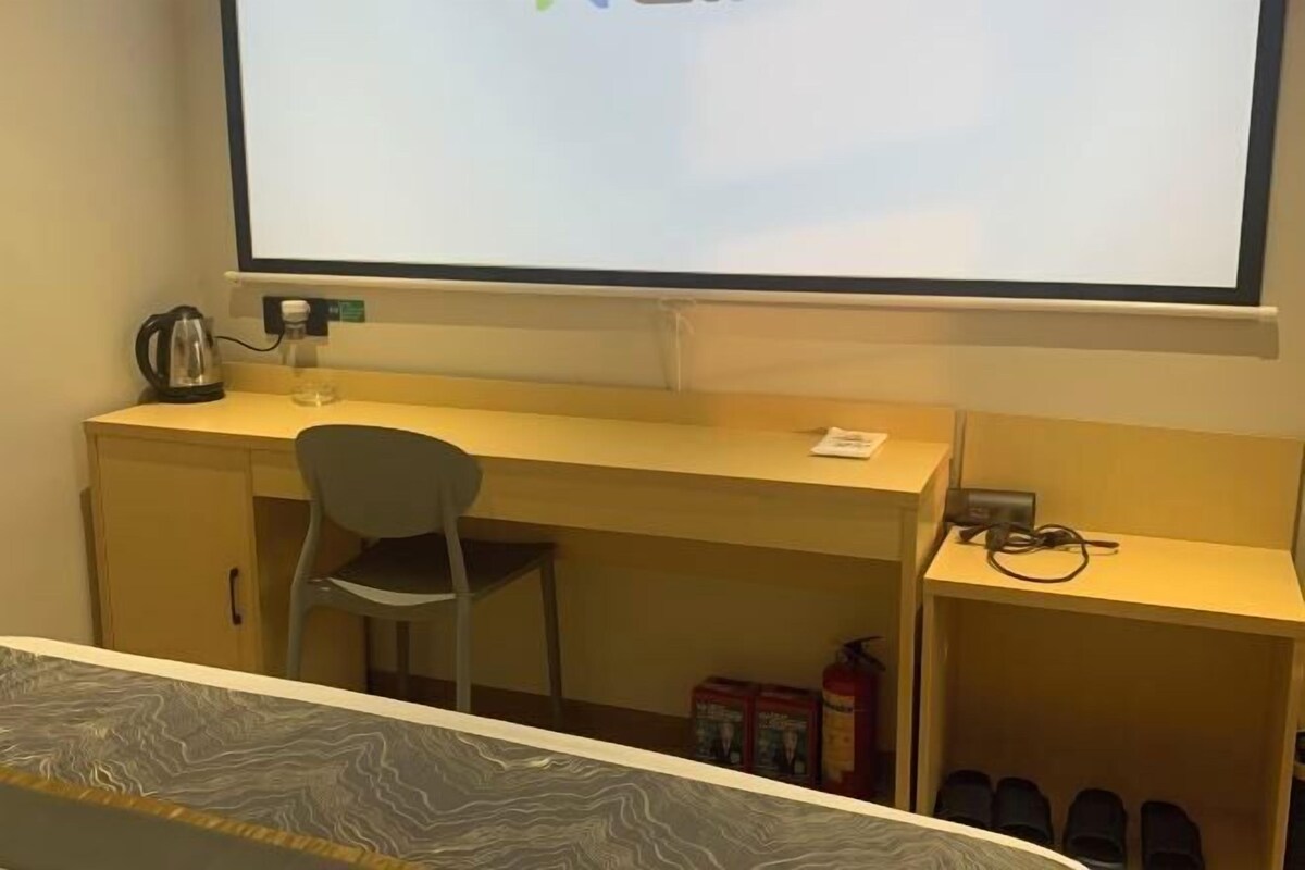 A room with a bed, a desk, a chair, a projector screen, a kettle, a fire extinguisher, and a shoe rack.