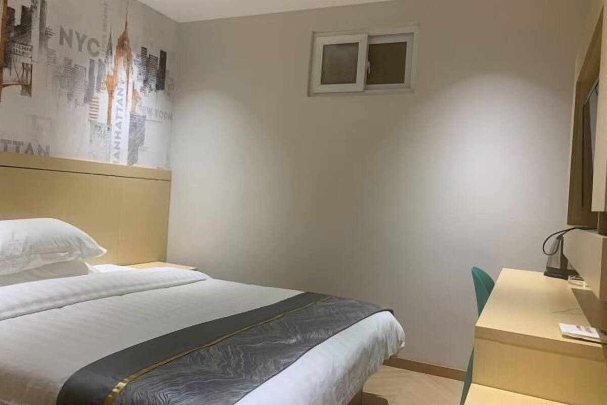 A hotel room with a bed, a desk, a chair, a window, and a wall with a cityscape mural.