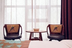 Habitación de hotel con dos sillas con cojines estampados, una mesita redonda con un frutero y una cama con colcha floral.