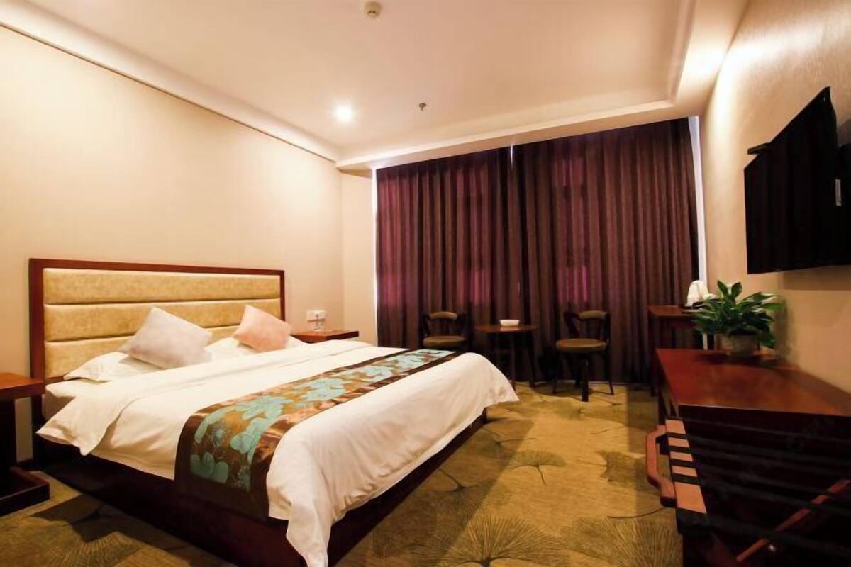 A hotel room with a bed, a desk, a chair, a small table, and a potted plant.