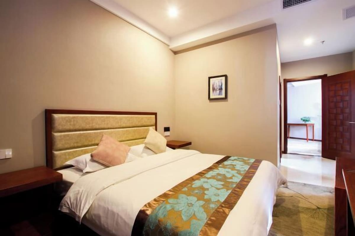 A hotel room with a bed, bedside tables, a nightstand, a framed picture on the wall, and a door leading to another room.