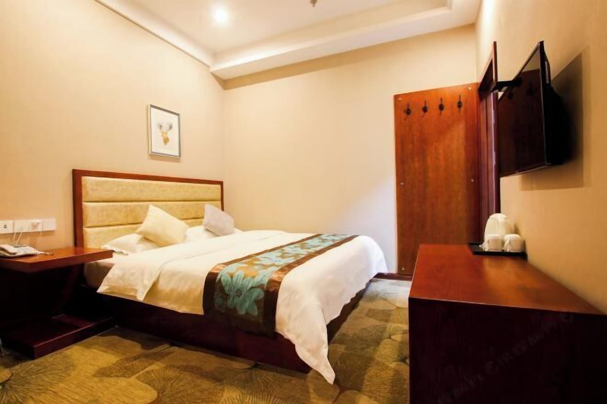 A hotel room with a bed, a wooden nightstand, a desk, a wardrobe, and a flat-screen TV mounted on the wall.