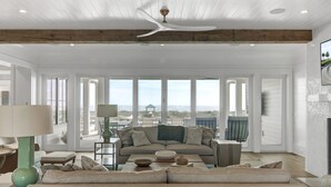 A spacious living room with a large window, a ceiling fan, and a view of the beach.