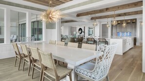 A spacious dining area with a long white table and wicker chairs, a kitchen with white cabinets, and a wooden ceiling.