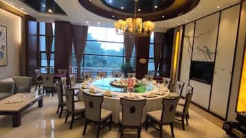 Uma sala de jantar com uma mesa redonda posta para seis pessoas, rodeada de cadeiras, com um lustre acima e vista para o exterior através de grandes janelas.